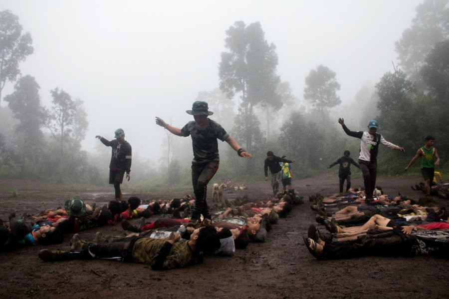 A group of young men are seen lying face up on the muddy ground in a forested area shrouded in mist. They appear to be undergoing military or survival training, with instructors walking over them, balancing on their bodies. The scene is intense and the environment is damp and foggy, adding to the sense of hardship.