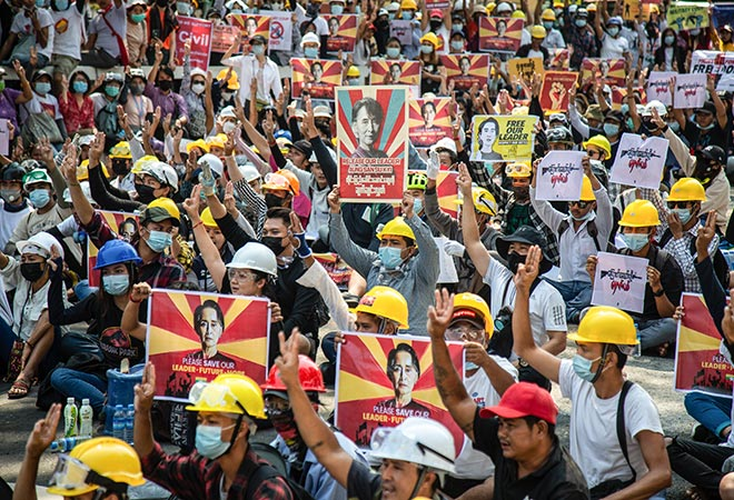 A large group of protesters in Myanmar facing the Burmese Crisis , wearing helmets and face masks, gather in a peaceful demonstration. Many hold up posters featuring the image of Aung San Suu Kyi, calling for her release and expressing support for democracy. The crowd is unified in raising three-finger salutes, a symbol of resistance.